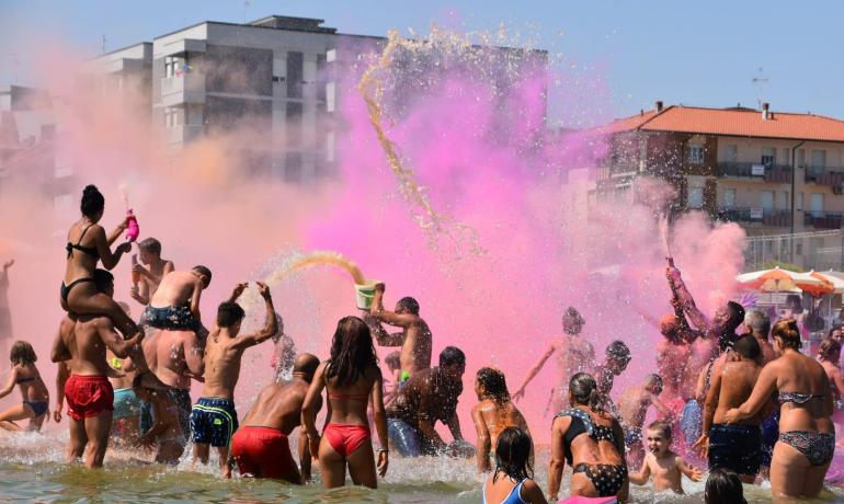 Persone festeggiano con colori in spiaggia durante una giornata soleggiata.