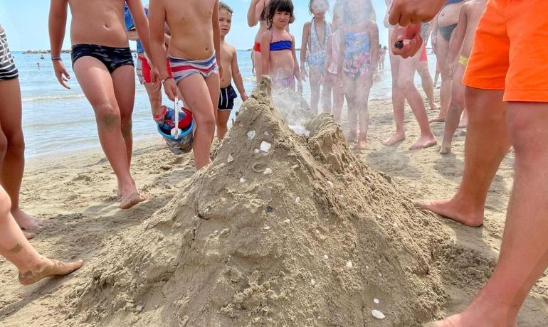 Bambini costruiscono un vulcano di sabbia sulla spiaggia, osservati da un adulto.