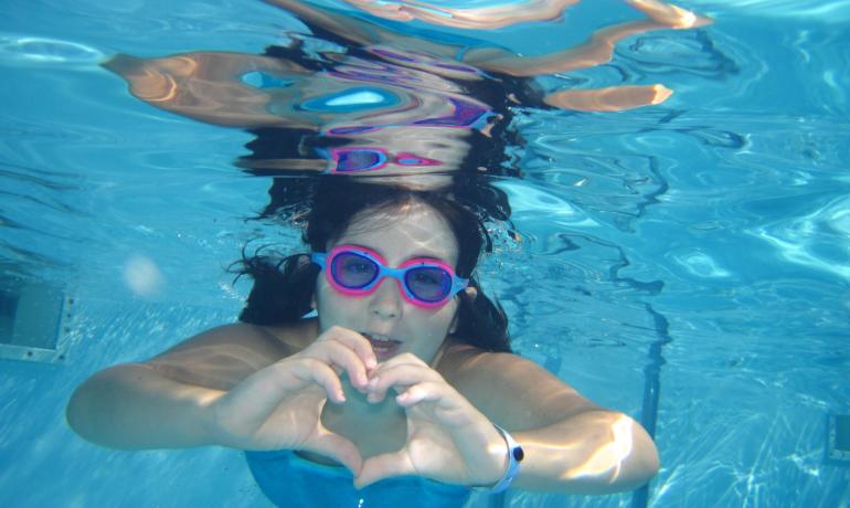Persona sott'acqua con occhiali da nuoto rosa fa un cuore con le mani.