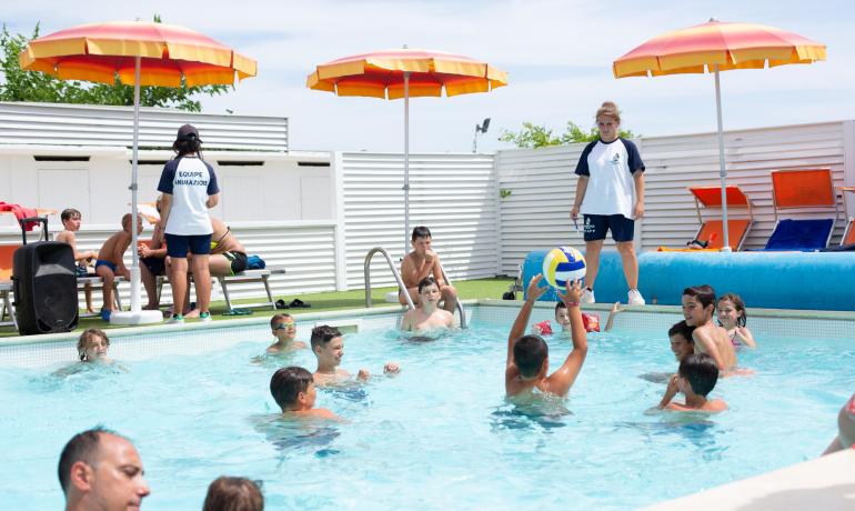 Bambini giocano felici in piscina, sotto ombrelloni colorati, sorvegliati da animatori.