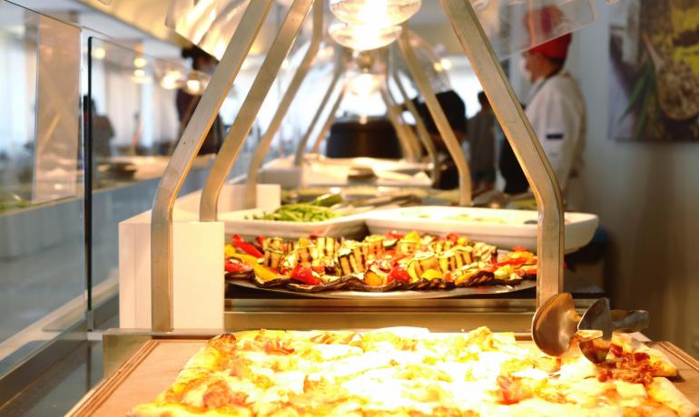 Buffet con pizza e verdure grigliate, chef sullo sfondo.