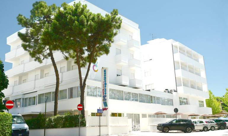Hotel moderno con balconi bianchi e parcheggio, circondato da alberi.