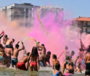 Persone festeggiano in spiaggia lanciando polvere colorata, creando un'atmosfera vivace e gioiosa.