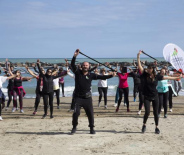 Gruppo energico si allena sulla spiaggia, sollevando bastoni, mare e cielo azzurro sullo sfondo.