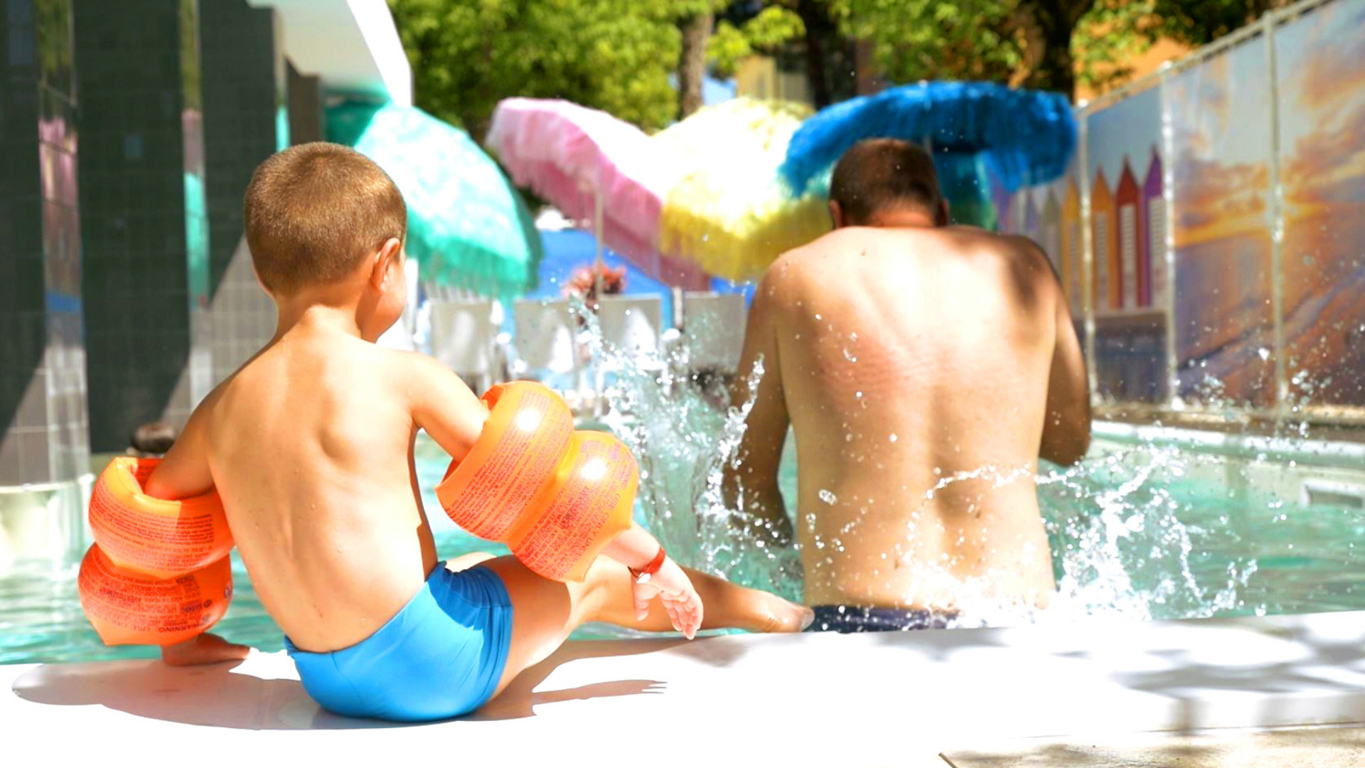 Bambino e uomo in piscina con braccioli, ombrelloni colorati sullo sfondo.