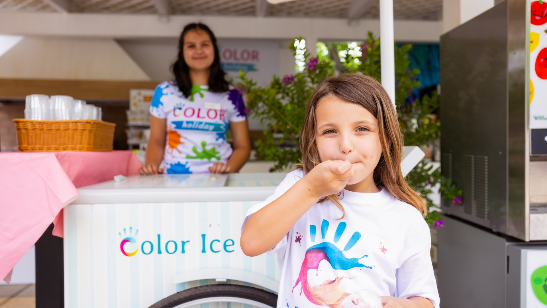 Bambina mangia gelato davanti a un chiosco Color Ice con una donna sorridente.