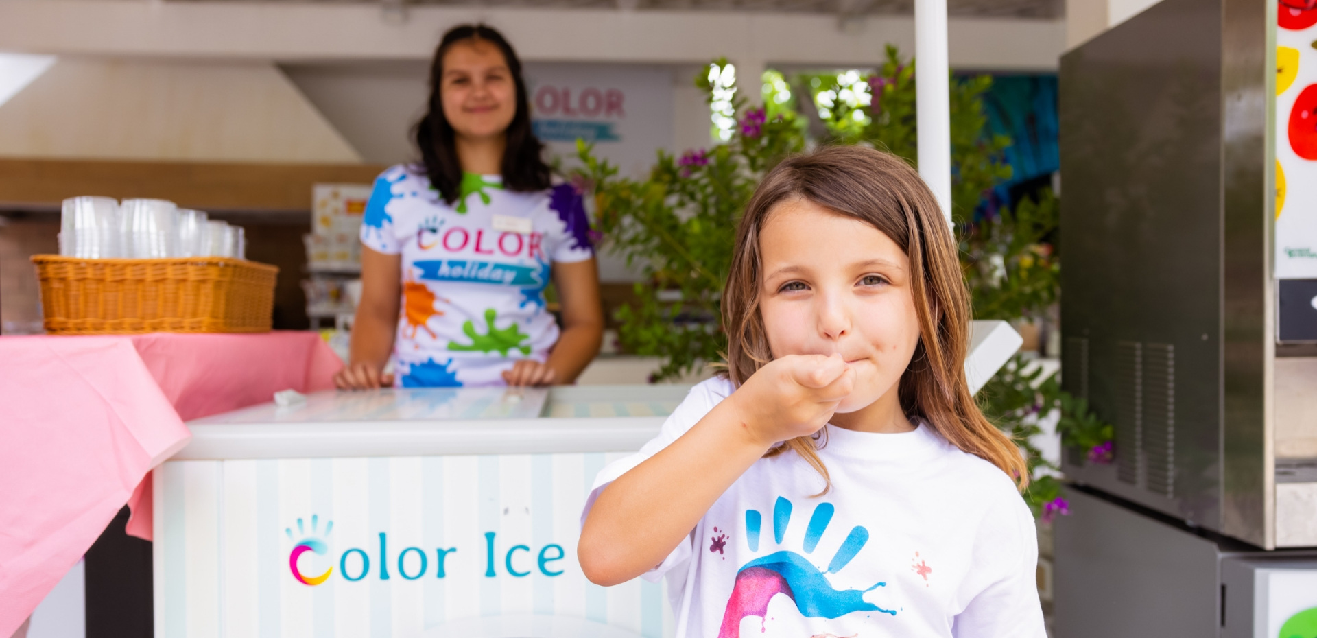 Bambina mangia gelato davanti a un chiosco colorato con una donna sorridente.