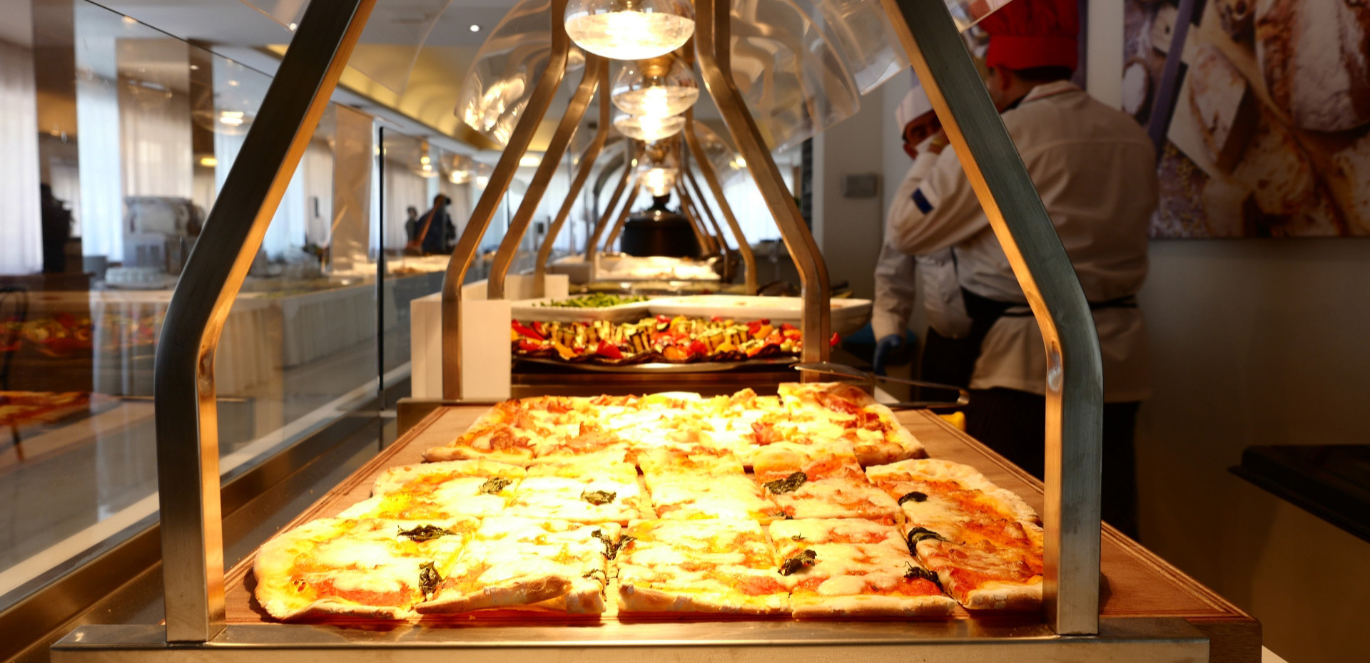 Buffet con pizza e verdure, chef sullo sfondo in cucina luminosa.