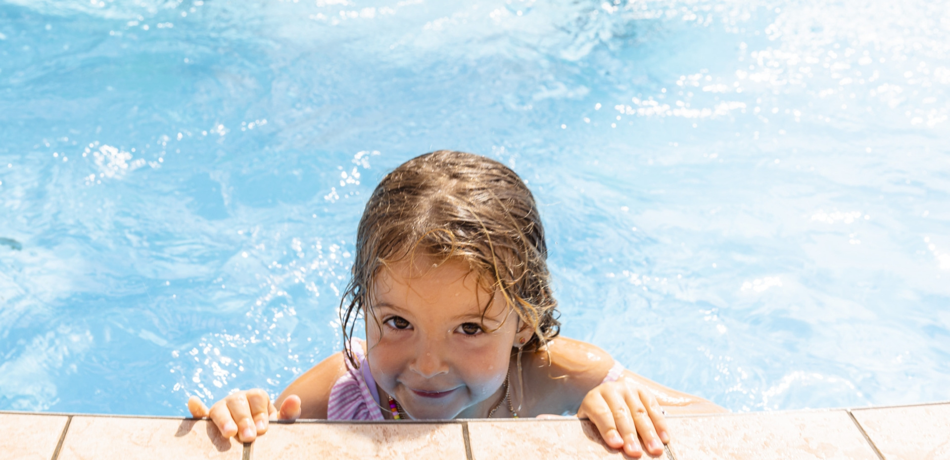 Bambina sorridente in piscina, si aggrappa al bordo sotto il sole estivo.