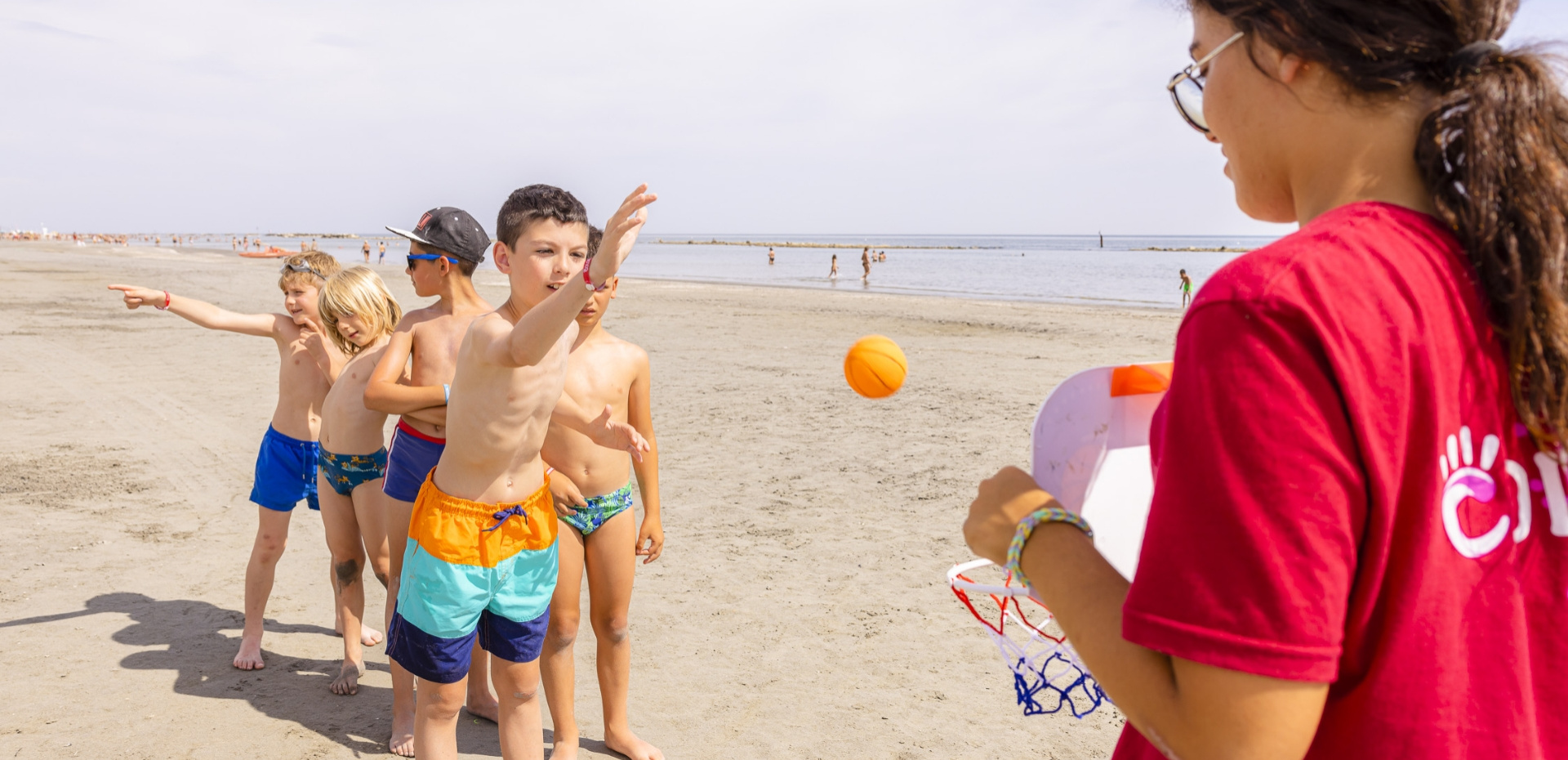 Bambini giocano a basket sulla spiaggia, guidati da un'istruttrice in maglietta rossa.
