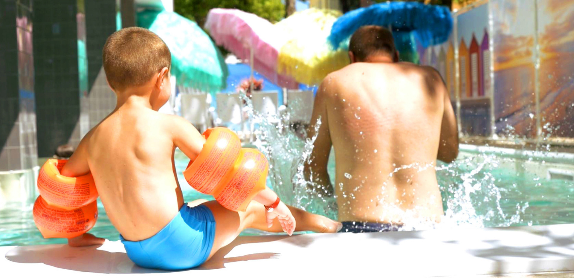 Bambino e uomo in piscina con braccioli, ombrelloni colorati sullo sfondo.