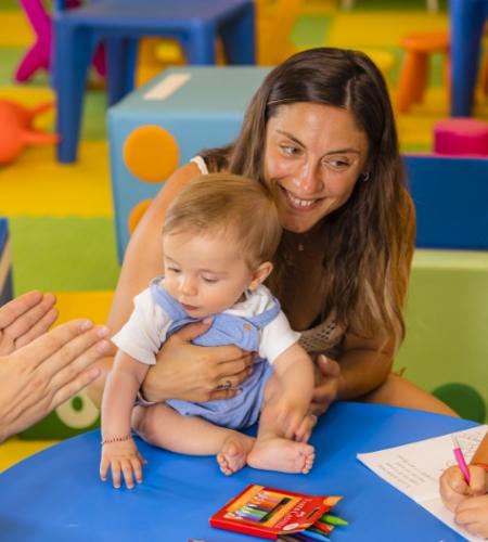 Bambino seduto al tavolo con colori, accompagnato da un adulto sorridente.