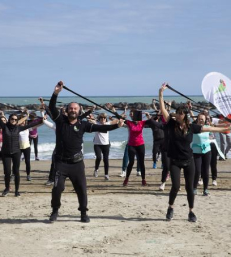 Gruppo energico si allena sulla spiaggia, sollevando bastoni, mare e cielo azzurro sullo sfondo.