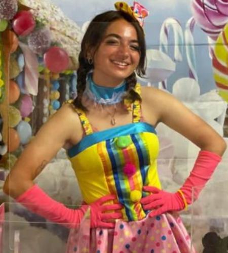 Persona in costume colorato, con guanti rosa, sfondo di dolci e caramelle.