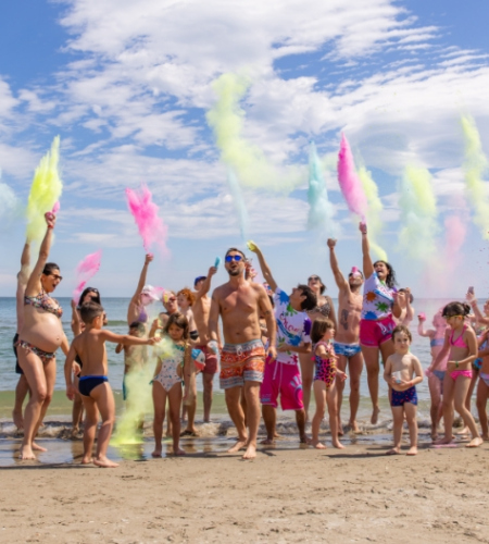Gruppo di persone in spiaggia lancia polveri colorate in aria, celebrando con gioia.