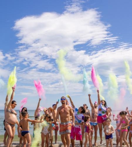 Gruppo festeggia in spiaggia lanciando polveri colorate sotto un cielo limpido.