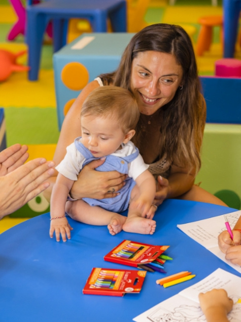Bambino seduto al tavolo con colori, accompagnato da un adulto sorridente.