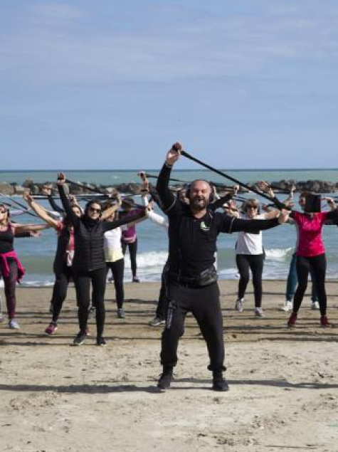 Gruppo energico si allena sulla spiaggia, sollevando bastoni, mare e cielo azzurro sullo sfondo.