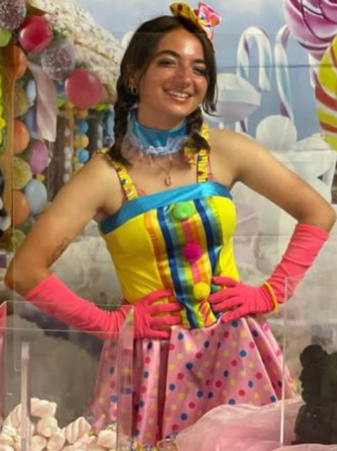 Persona in costume colorato, con guanti rosa, sfondo di dolci e caramelle.