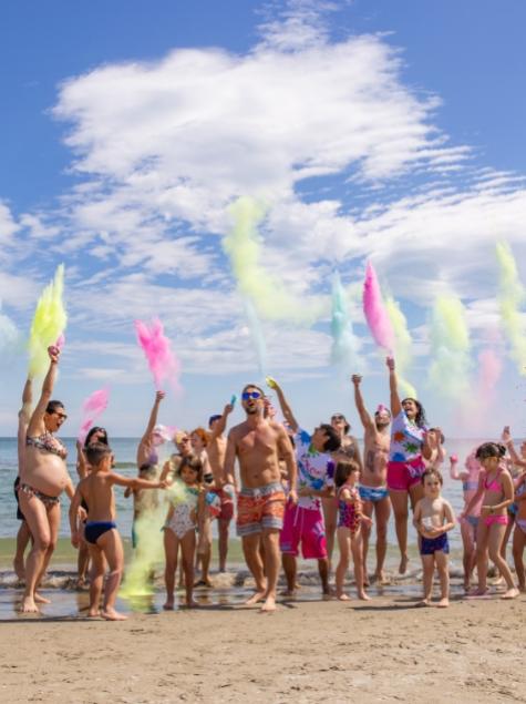 Gruppo di persone in spiaggia lancia polveri colorate in aria, celebrando con gioia.