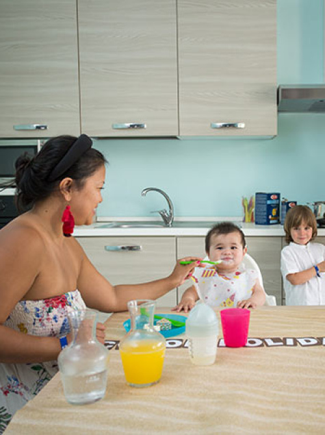 Famiglia in cucina, madre prepara, bambino sorride, altra donna dà da mangiare al piccolo.