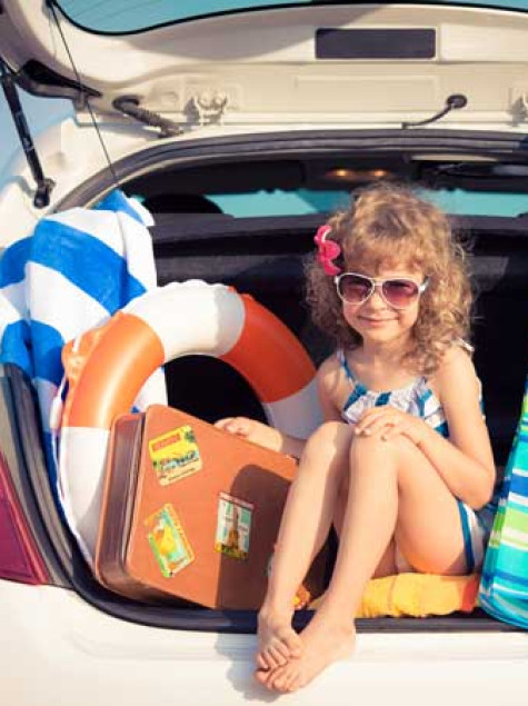 Bambina felice in auto con valigia, ombrellone e salvagente al mare.