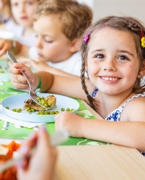 Bambina sorridente mangia un panino, con altri bambini sullo sfondo.