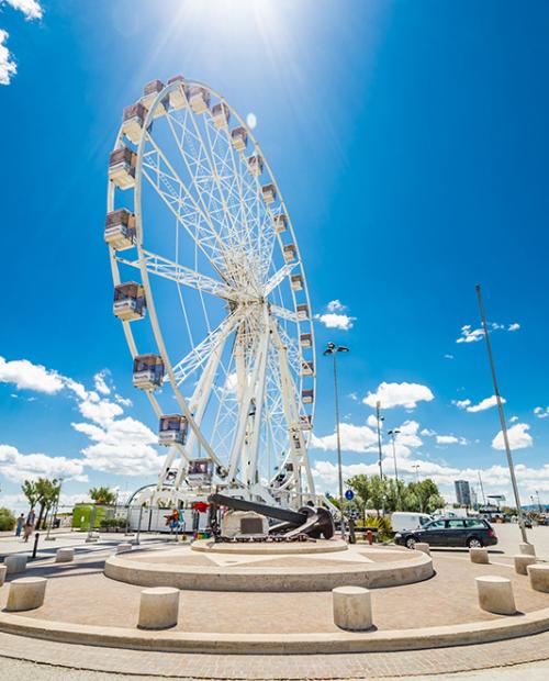 Ruota panoramica sul lungomare sotto un cielo blu con nuvole bianche.