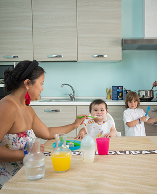 Famiglia in cucina, madre prepara, bambino sorride, altra donna dà da mangiare al piccolo.