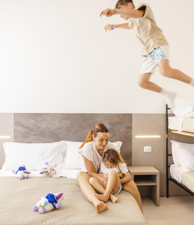 Madre abbraccia figlia sul letto, mentre un bambino salta dalla cuccetta.