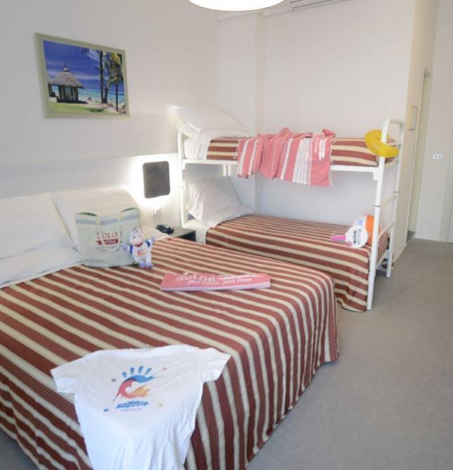 Camera d'hotel con letto matrimoniale e a castello, decorazioni colorate e valigia rossa.